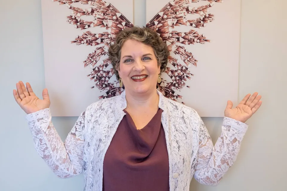 Dra. Patrícia de braços abertos diante de dois quadros que juntos formam uma borboleta feita de flores secas em tons de coral.