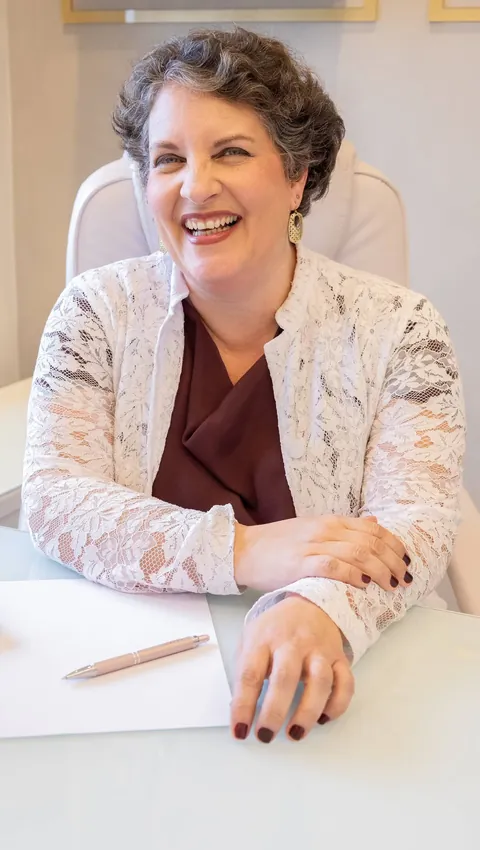 Dra. Patrícia, de blusa vinho e casaco de renda branca, ri sentada à mesa de atendimento.