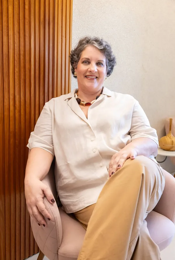 Dra. Patrícia Barros sentada numa poltrona, de camisa clara e calça caramelo, diante de um painel de madeira.